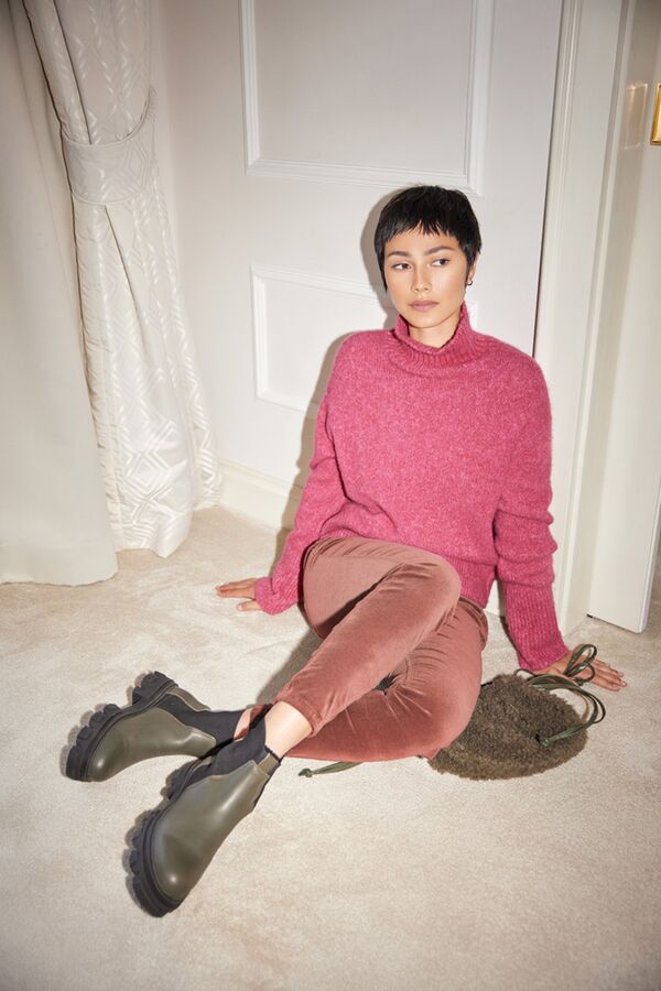 Weibliches Modell in pinken Turtleneck-Pullover und rosefarbener Skinny-Jeans von Luisa-Cerano posiert sitzend auf dem Boden