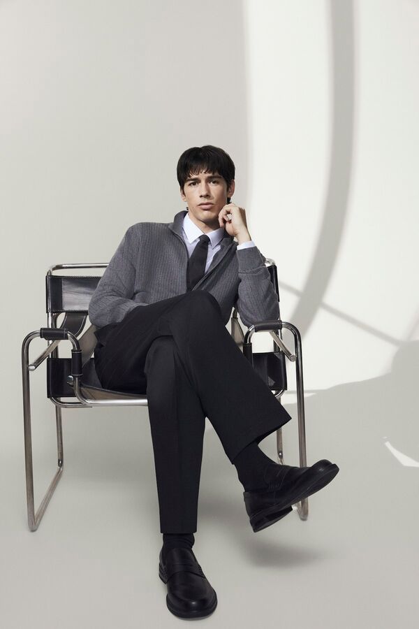 Männliches Modell in schwarzer Hose und grauen Mantel von BOSS posiert auf einem Stuhl