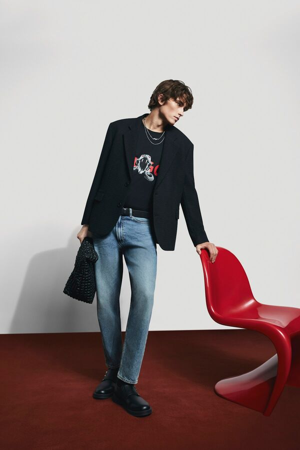 Männliches Modell in blauer Denim, schwarzem Shirt und schwarzen Sakko von BOSS lehnt sich an einen roten Designer-Stuhl