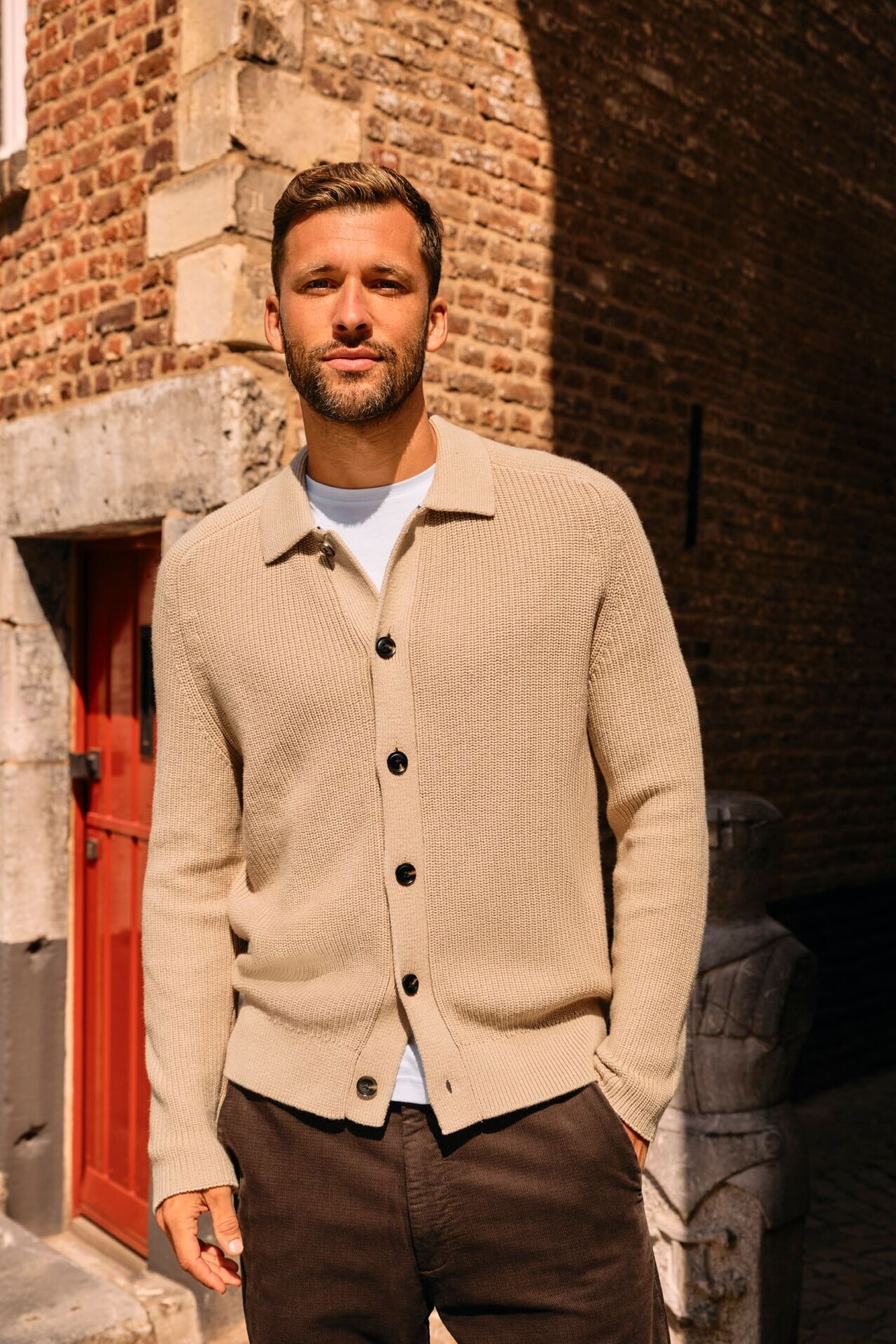 Männliches Modell in dunkelbrauner Hose und hellbeigen Strick-Cardigan posiert lässig vor einem Haus