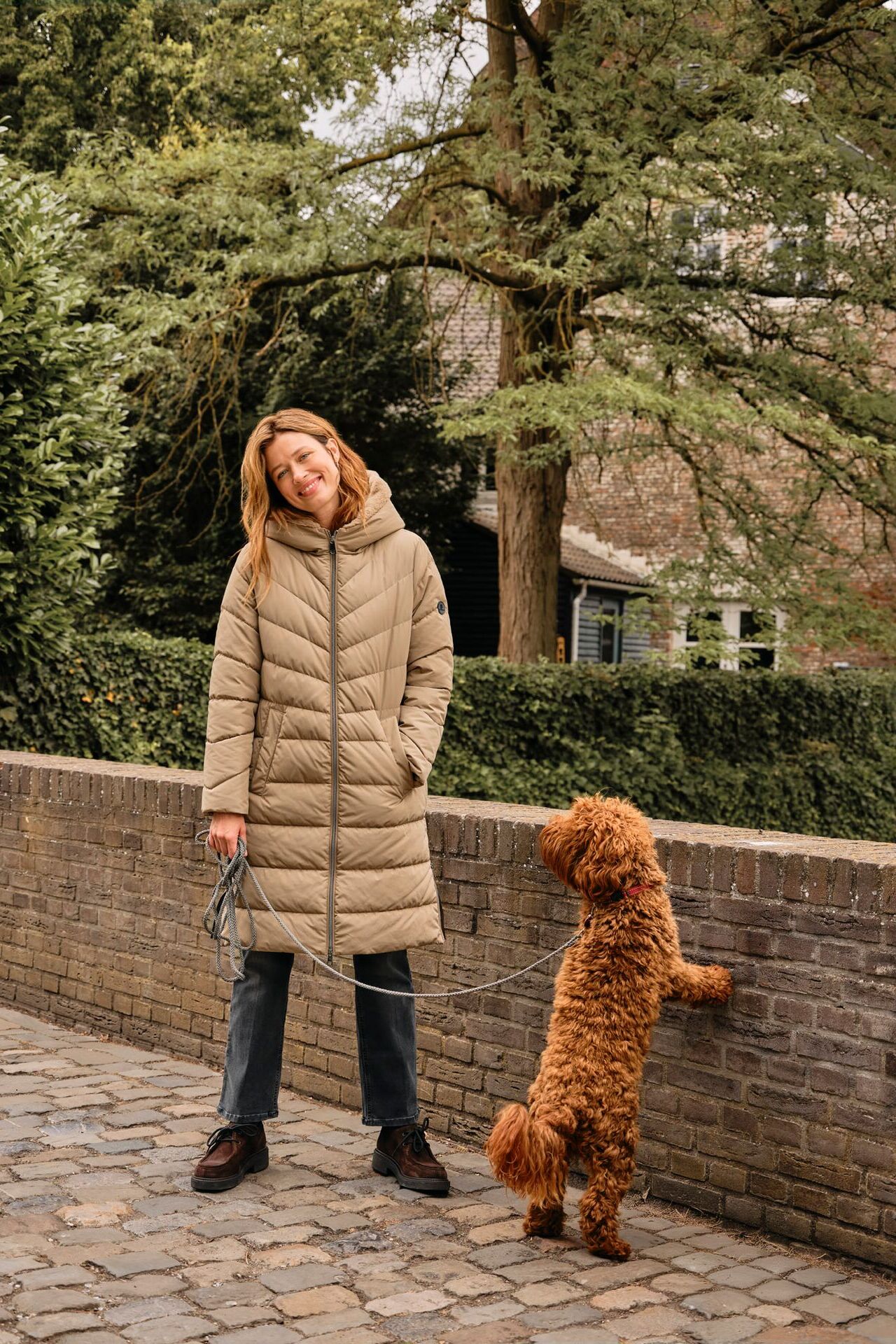 Weibliches Modell in hellbeigen Steppmantel geht mit einem süßen Hund Gassi