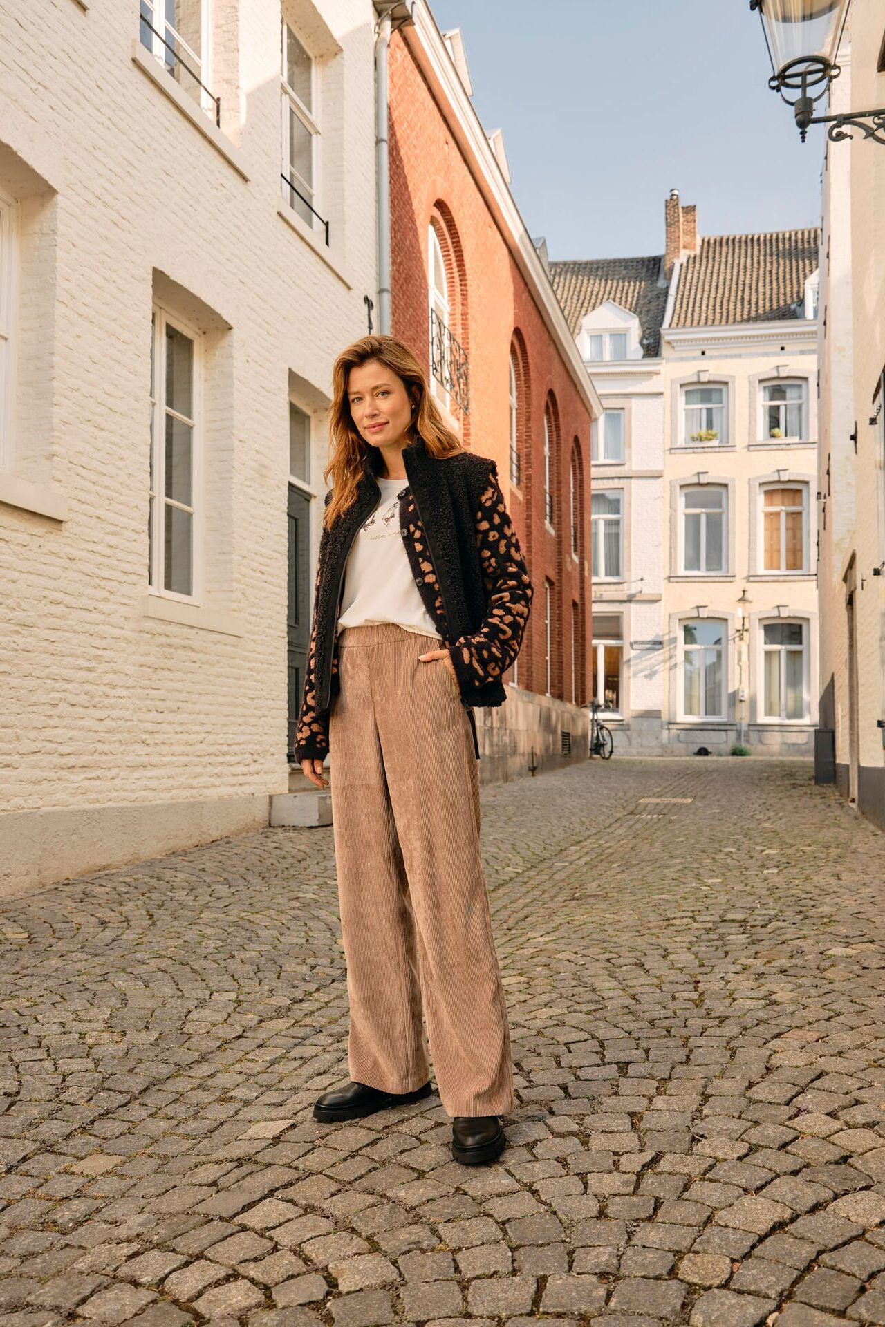 Weibliches Modell in hellbrauner Cordhose, weißen Shirt und gemusterten Jacke posiert in einer Gasse