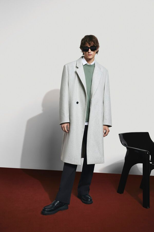 Männliches Modell in schwarzer Anzughose, hellgrünem Pullover und langen weißen Trenchcoat von BOSS posiert cool mit einer Sonnenbrille