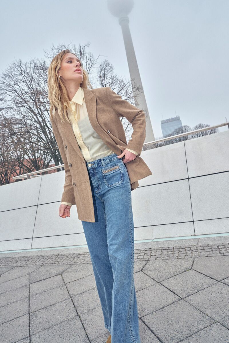 Weibliches Modell in blauer Denim-Jeans, hellgelber Bluse und braunem Business-Blazer blickt zur Seite