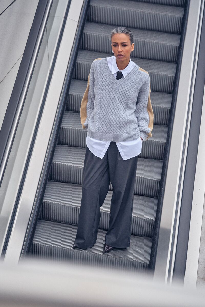 Weibliches Modell in Dunkelgrauer Businesshose, weißer Bluse und grauen Strickpullover steht auf einer Rolltreppe