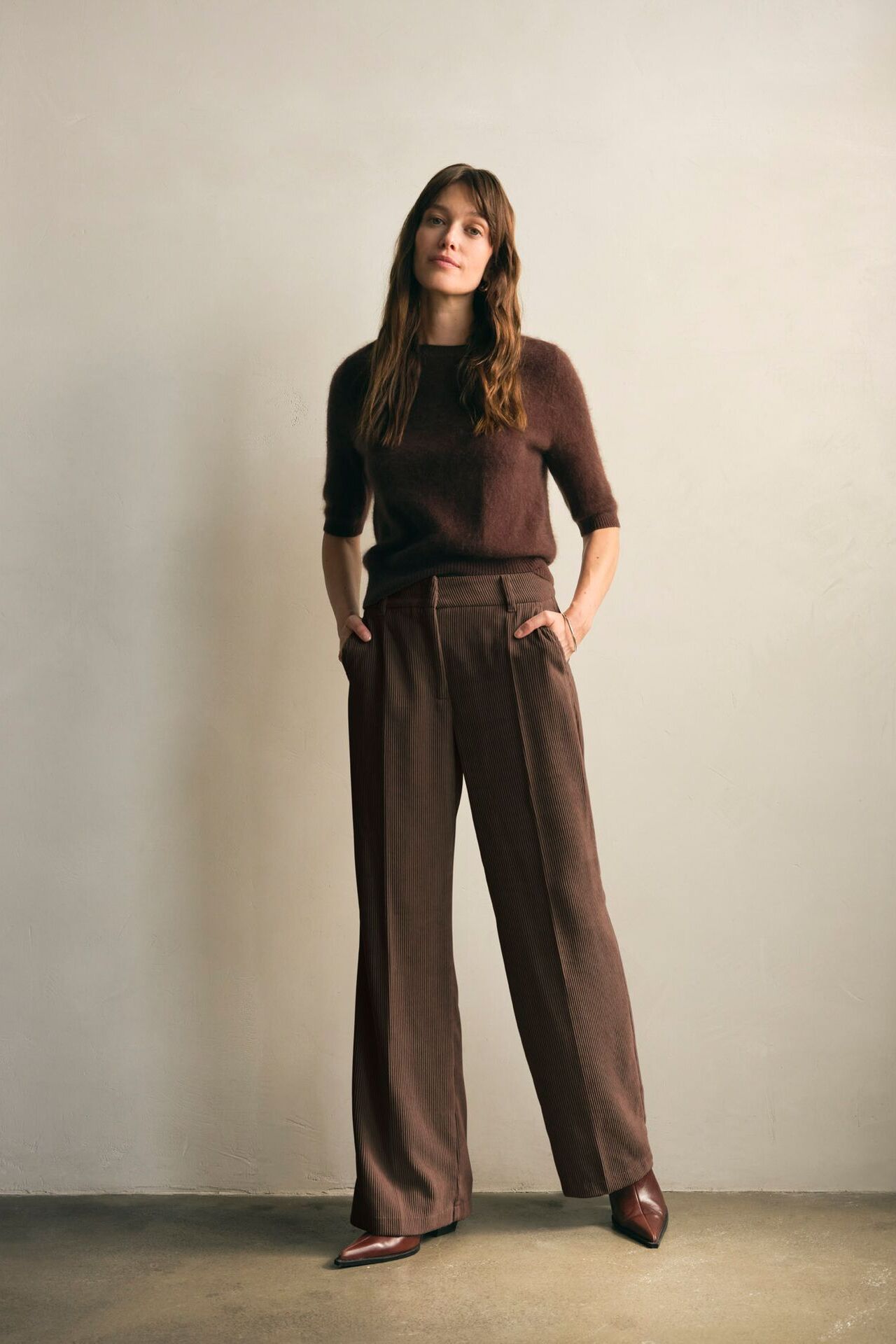 Weibliches Modell in Brauner Business-Hose und dunkelbraunen Strickpullover posiert mit den Händen in der Hosentasche