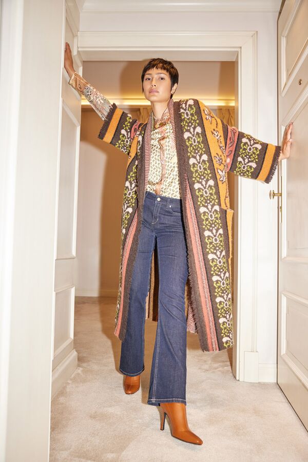 Weibliches Modell in Bluse mit Muster, gemusterten Mantel und blauer Jeans von Marc Cain posiert in einem Flur