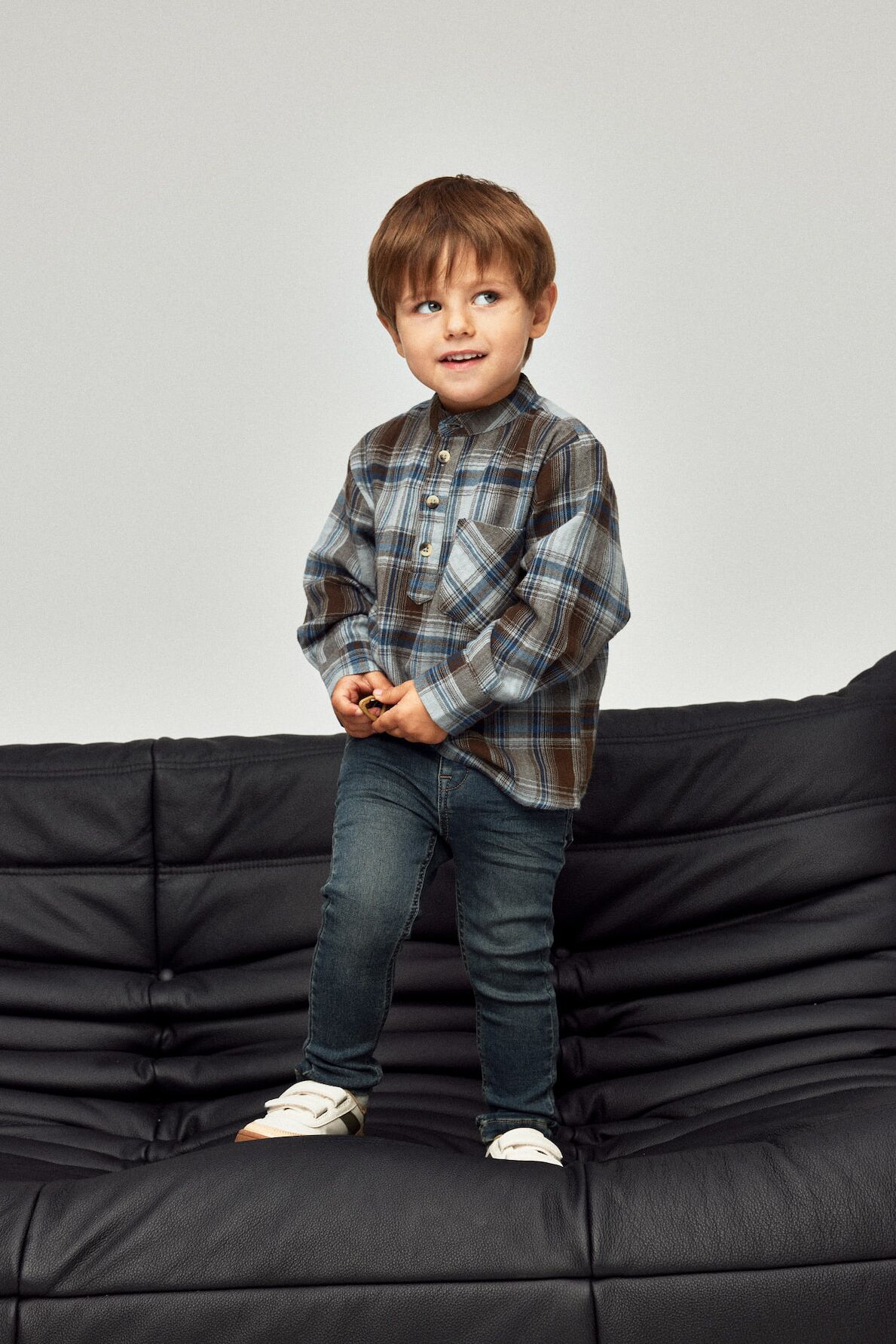Kleiner Junge in Jeans und Hemd von name it steht auf einem Sofa