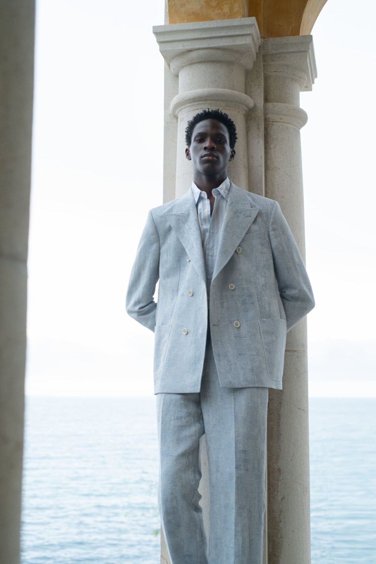 Männliches Modell in schicken hellgrauen Anzug von Baldessarini posiert an einer Steinsäule vor dem Meer