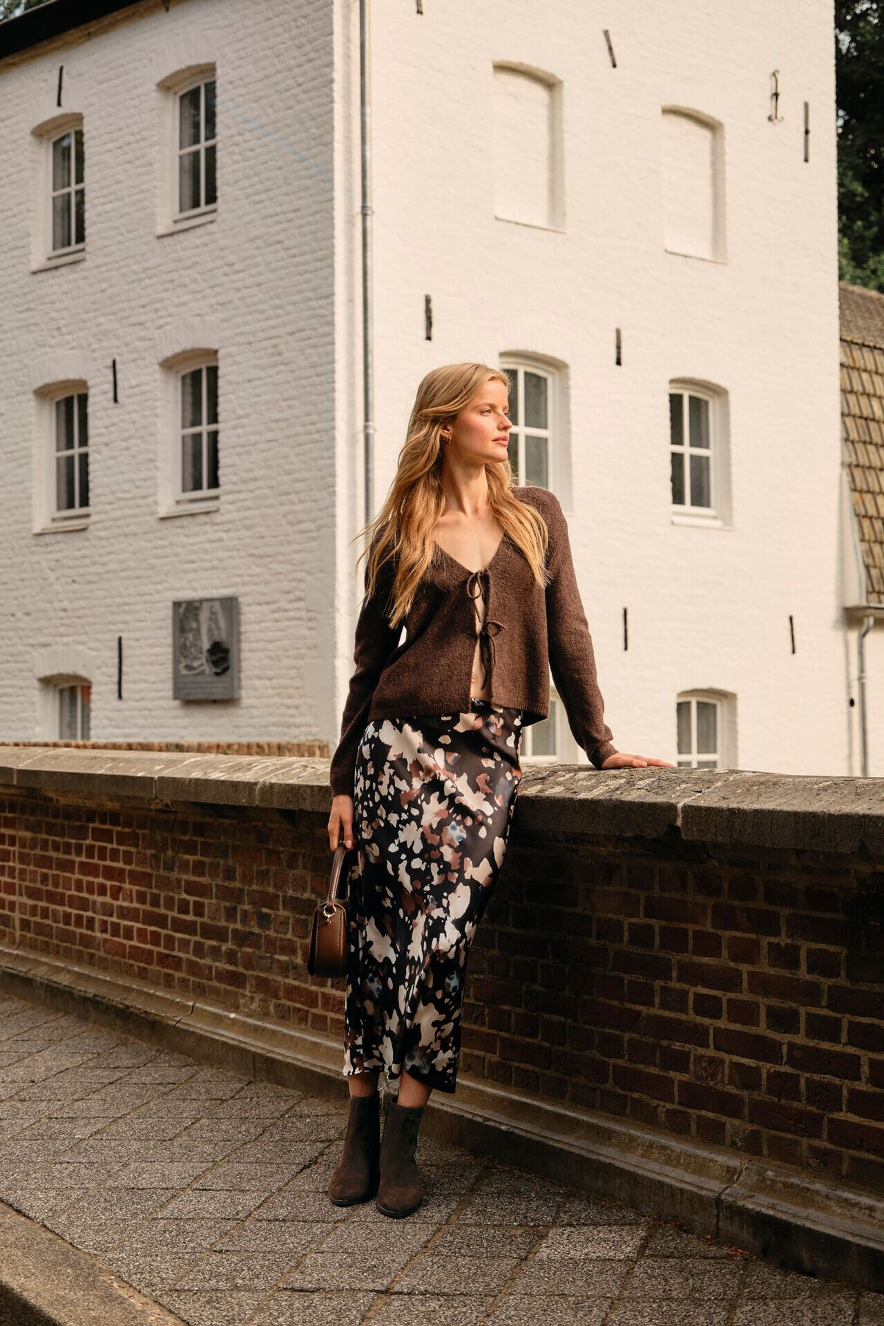 Weibliches Modell in angesagten Trend-Look aus Maxikleid und Cardigan posiert an einer Mauer