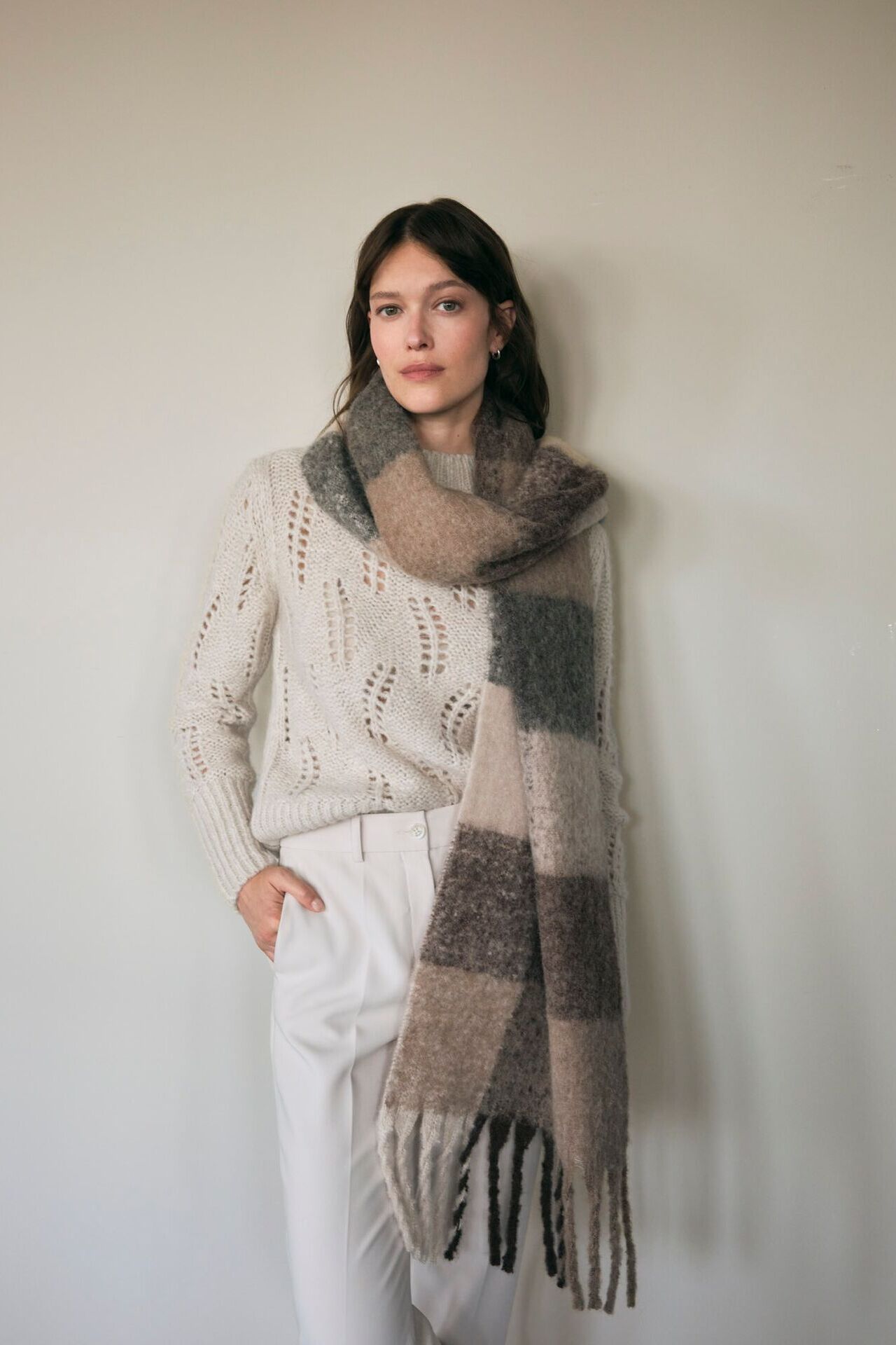 Weibliches Modell in weißer Hose, beigen Strickpullover und karierten XXL Schal posiert vor einer Wand