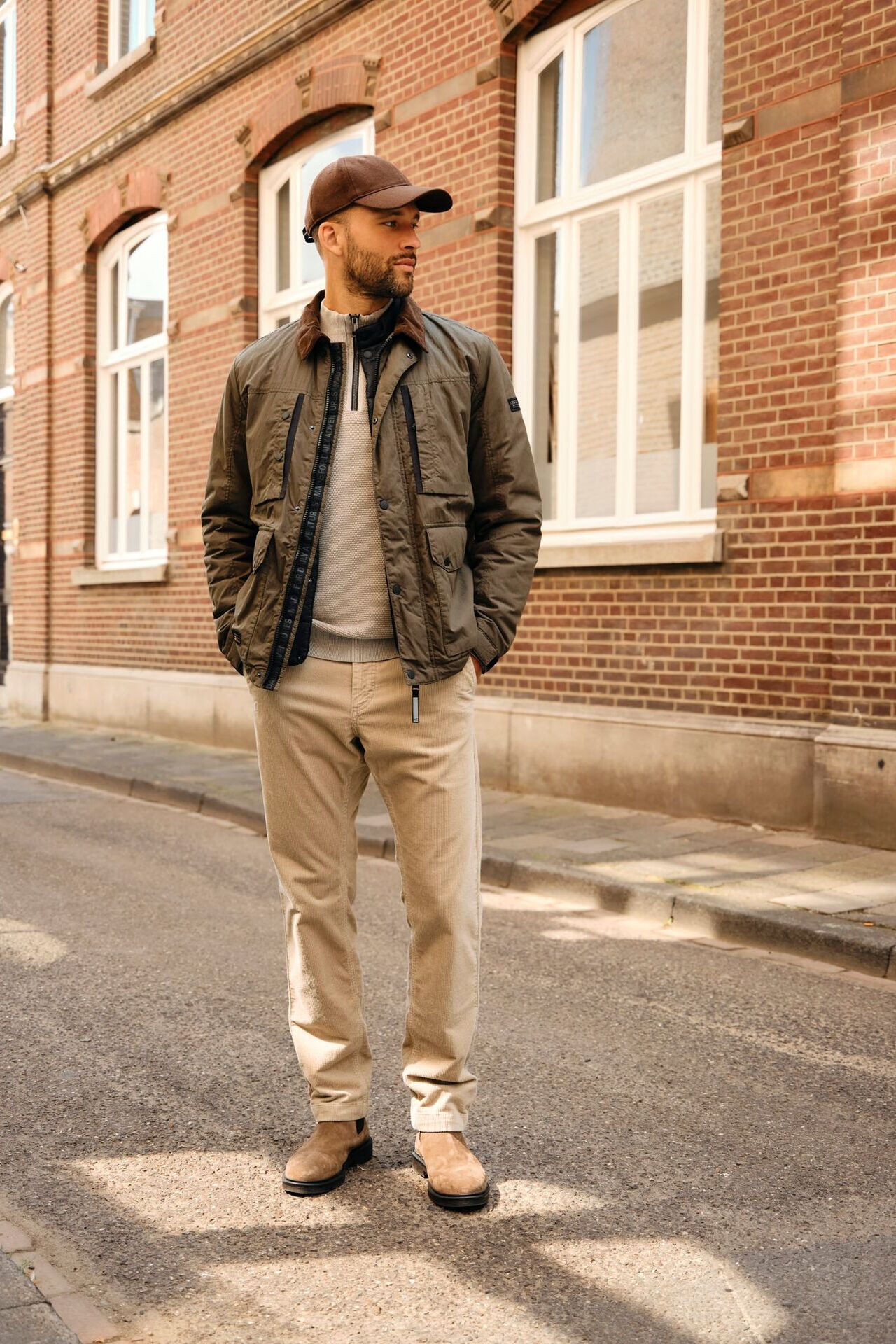 Männliches Modell in beiger Chinohose, grauen Troyer und gruener ÜBergangsjacke steht auf einer Straße