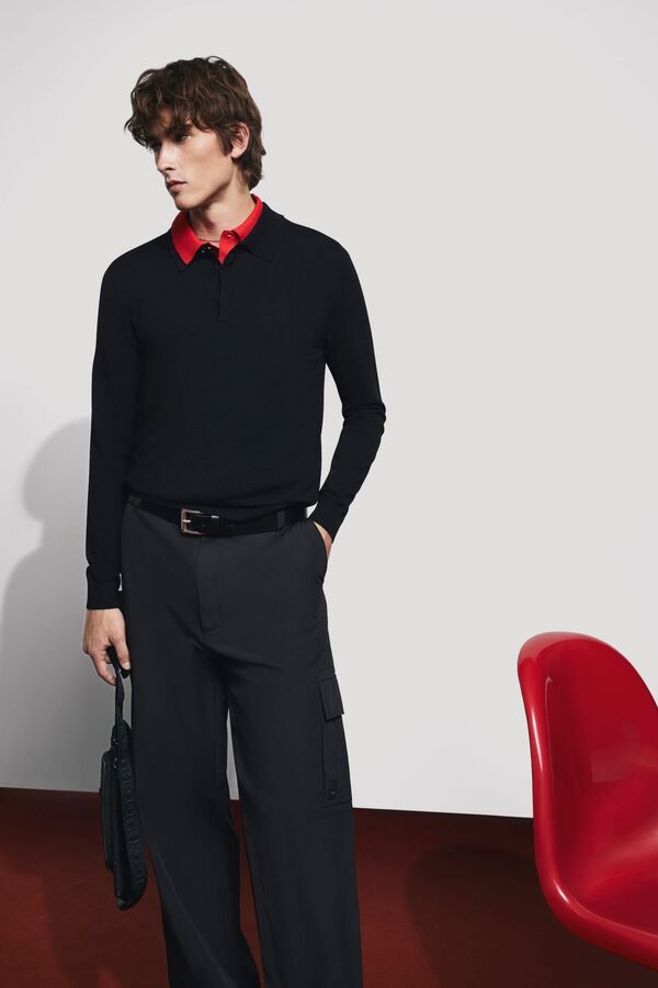 Männliches Modell mit schwarzer Anzughose, rotem Hemd und schwarzen Pullover von HUGO posiert neben einem roten Stuhl