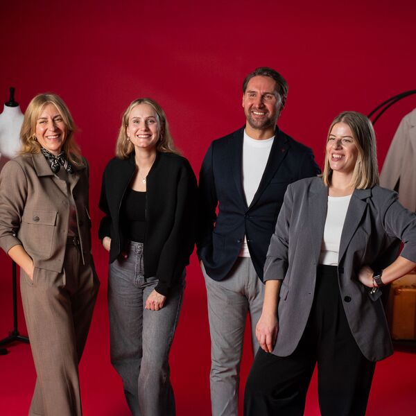 Drei Frauen und ein Mann stehen lachend vor rotem Hintergrund, alle in modischer Businesskleidung.