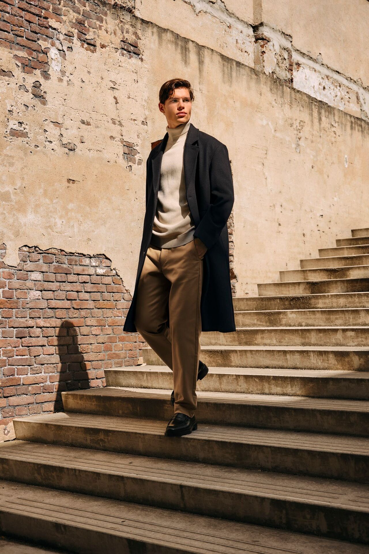 Männliches Modell in schicken Herbst-Look aus Chinohose, Rollkragenpullover und Mantel geht eine Treppe hinunter
