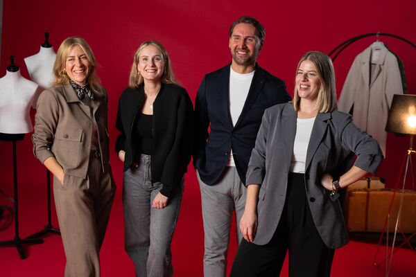 Drei Frauen und ein Mann stehen lachend vor rotem Hintergrund, alle in modischer Businesskleidung.