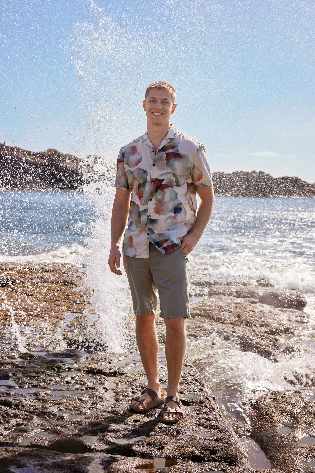 Männliches Modell in kurzer Hose und gemusterten Kurzarm-Hemd posiert lässig am Strand während Wellen einbrechen