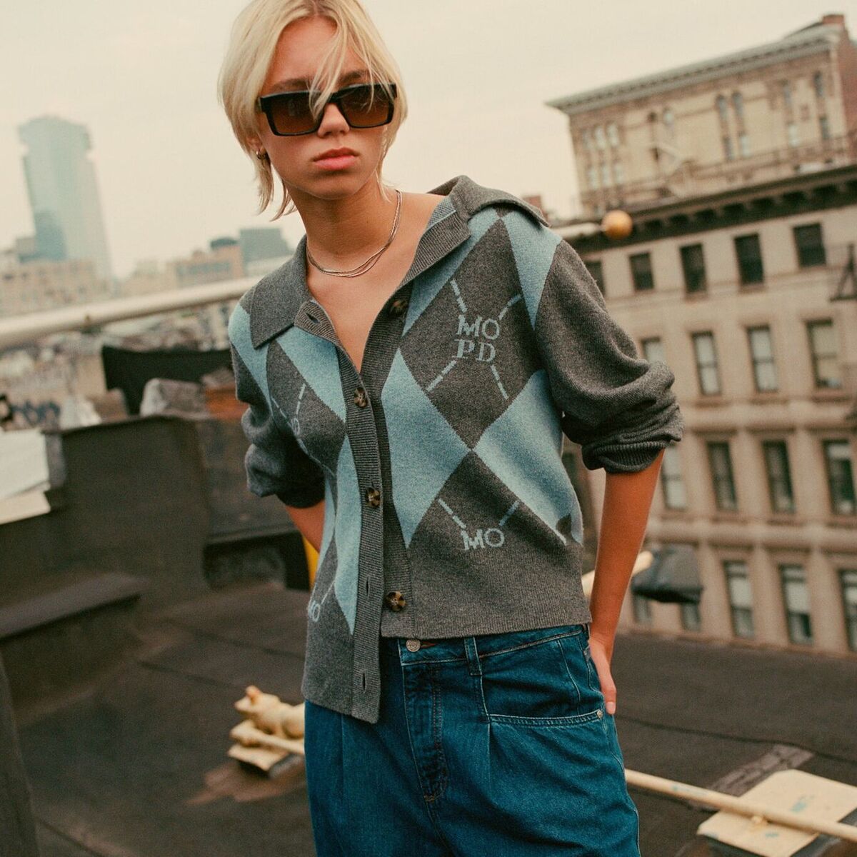 Weibliches Model in blauer Ballon-Jeans und gemusterten Strickjacke von Marc O Polo Denim auf einem Hochhausdach