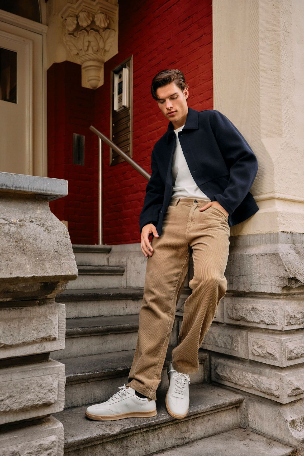 Männliches Modell in lässigem Herbst-Outfit aus Chinohose, T-Shirt und Übergangsjacke posiert cool vor einem Hauseingang
