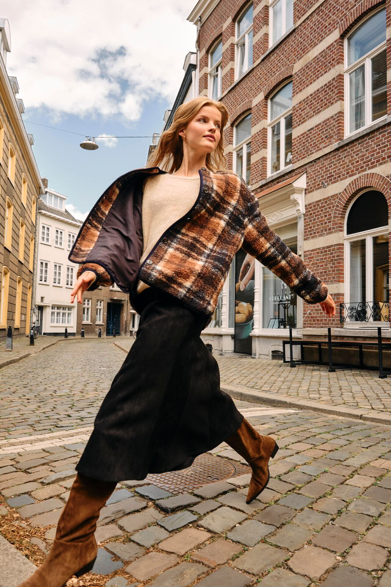 Weibliches Modell in schwarzen Maxirock, beigen Strickpullover und karierter Strickjacke läuft über eine gepflasterten Weg