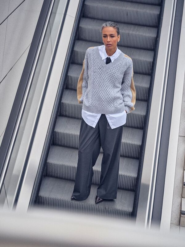 Weibliches Modell in Dunkelgrauer Businesshose, weißer Bluse und grauen Strickpullover steht auf einer Rolltreppe