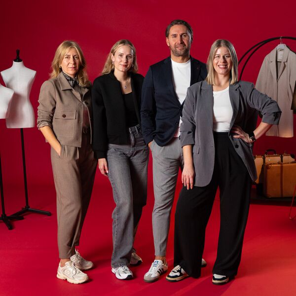 Drei Frauen und ein Mann stehen lachend vor rotem Hintergrund, alle in modischer Businesskleidung.