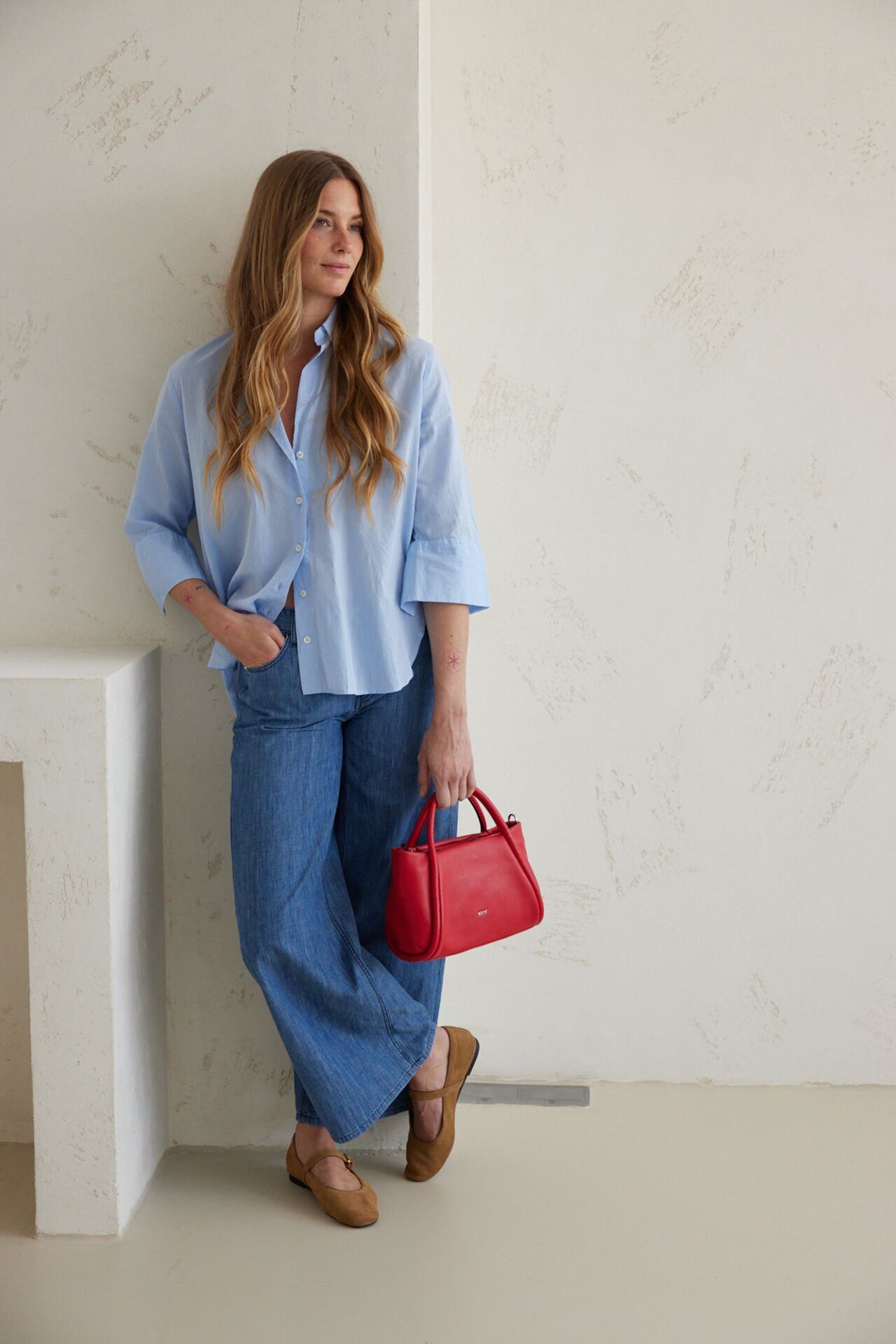Weibliches Modell in blauen Rock, hellblauer Bluse und einer roten Handtasche lehnt sich lässig an eine Wand