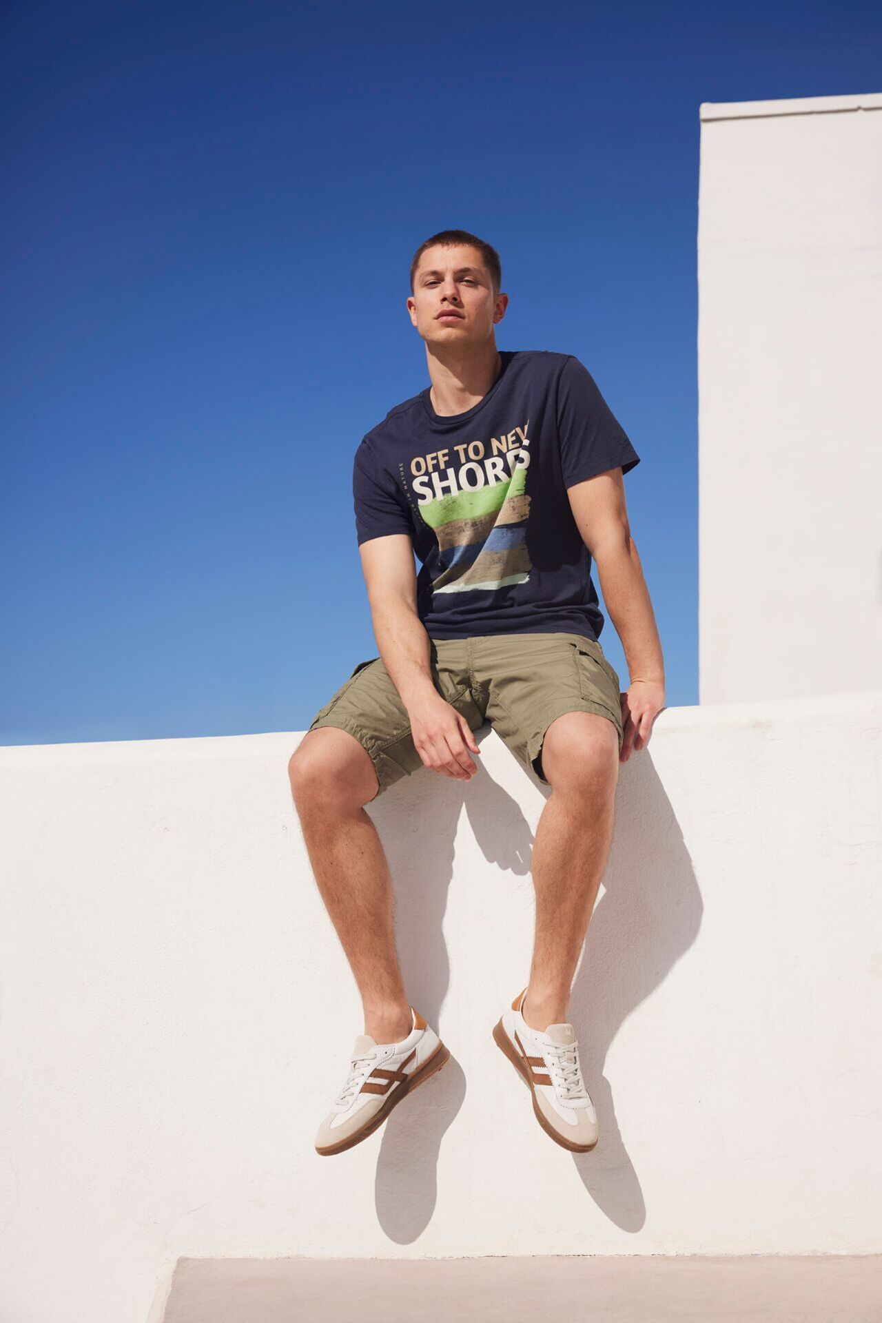 Männliches Modell in grüner Cargo-Shorts und dunkelblauen T-Shirt von Camel Active sitzt auf einer Mauer