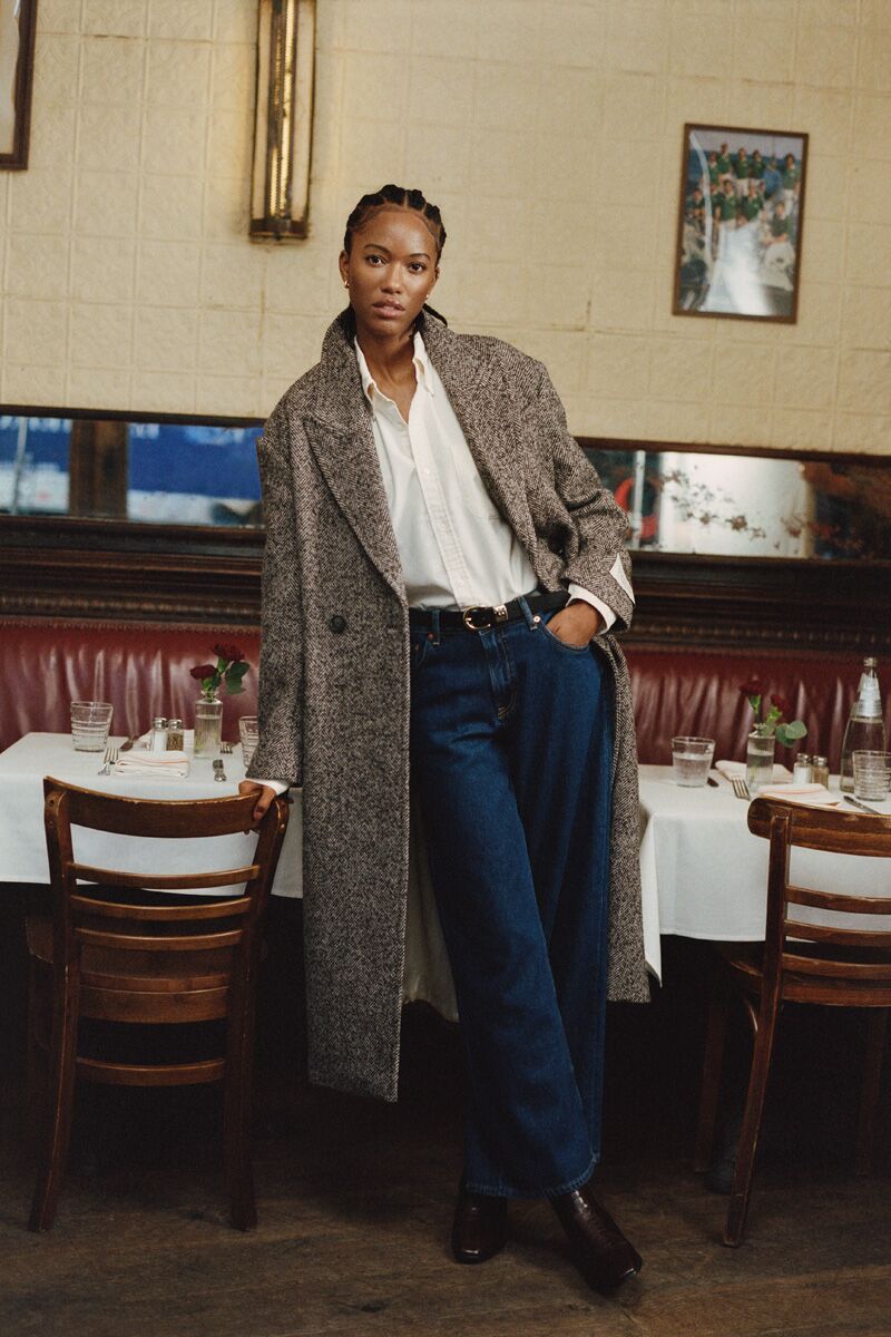 Weibliches Modell in dunkelblauer Jeans, weißer Bluse und melierten Mantel von GANT lehnt sich an einen Stuhl
