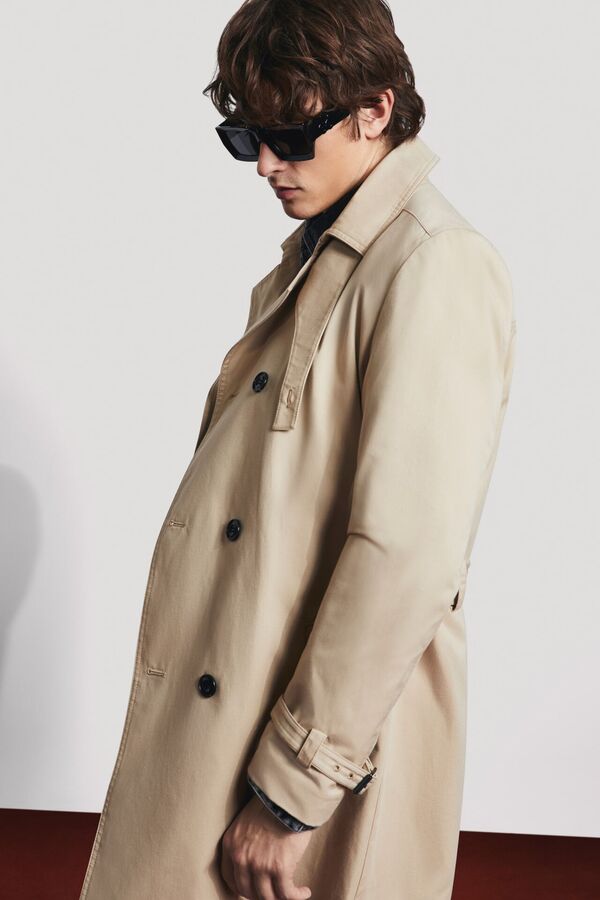 Männliches Modell in einem beigen Trenchcoat und einer schwarzen Sonnenbrille von BOSS RED blickt lässig auf den Boden