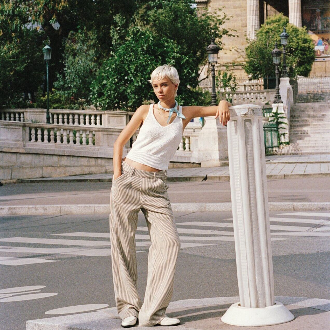 Weibliches Modell in beiger Joggerpants und weißen Crop-Top von Marc O'Polo lehnt sich an einen Steinpfeiler