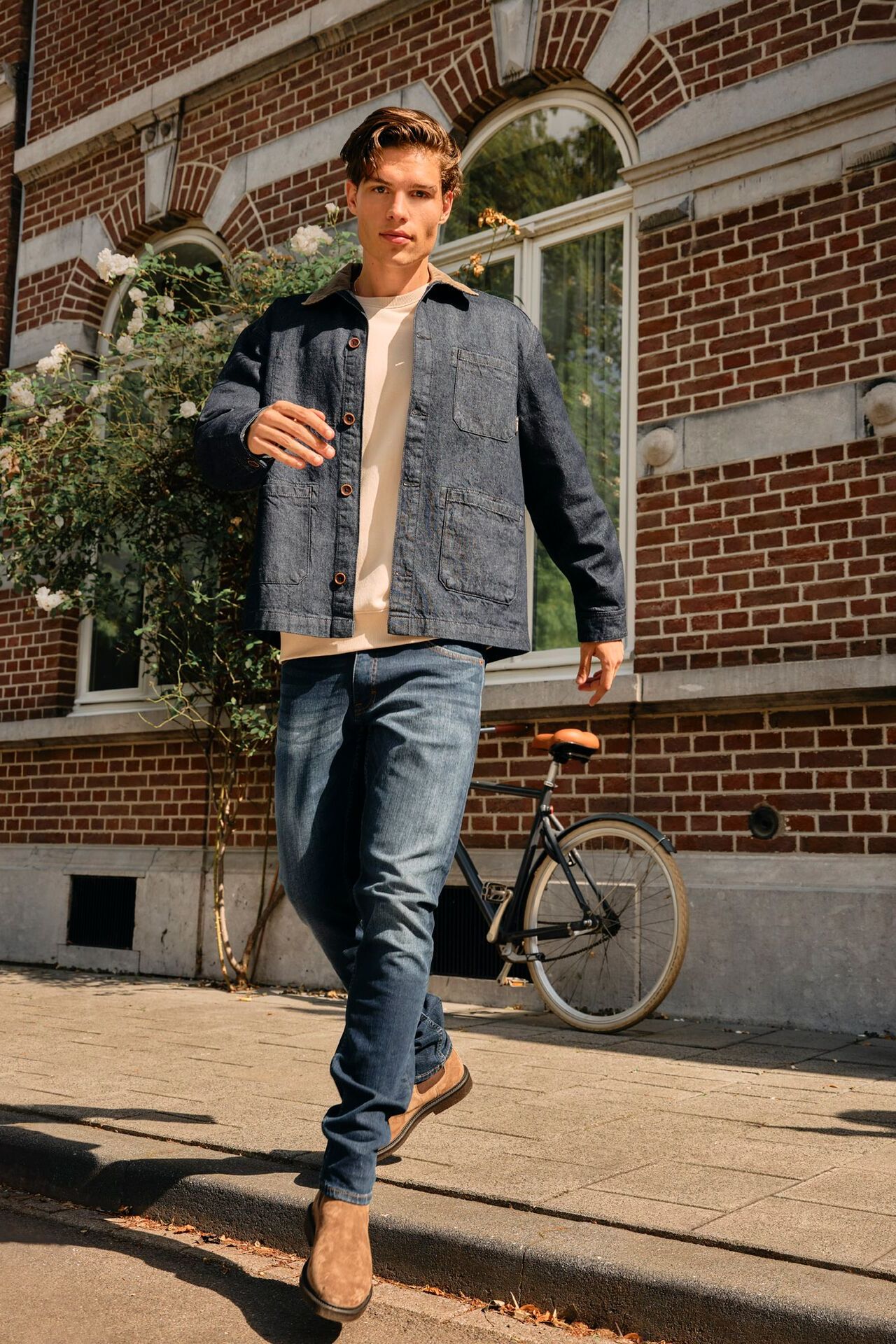 Männliches Modell in dunkelblauer Jeans, beigen Pullover und Graublauer Jeansjacke läuft vom Gehweg auf die Straße