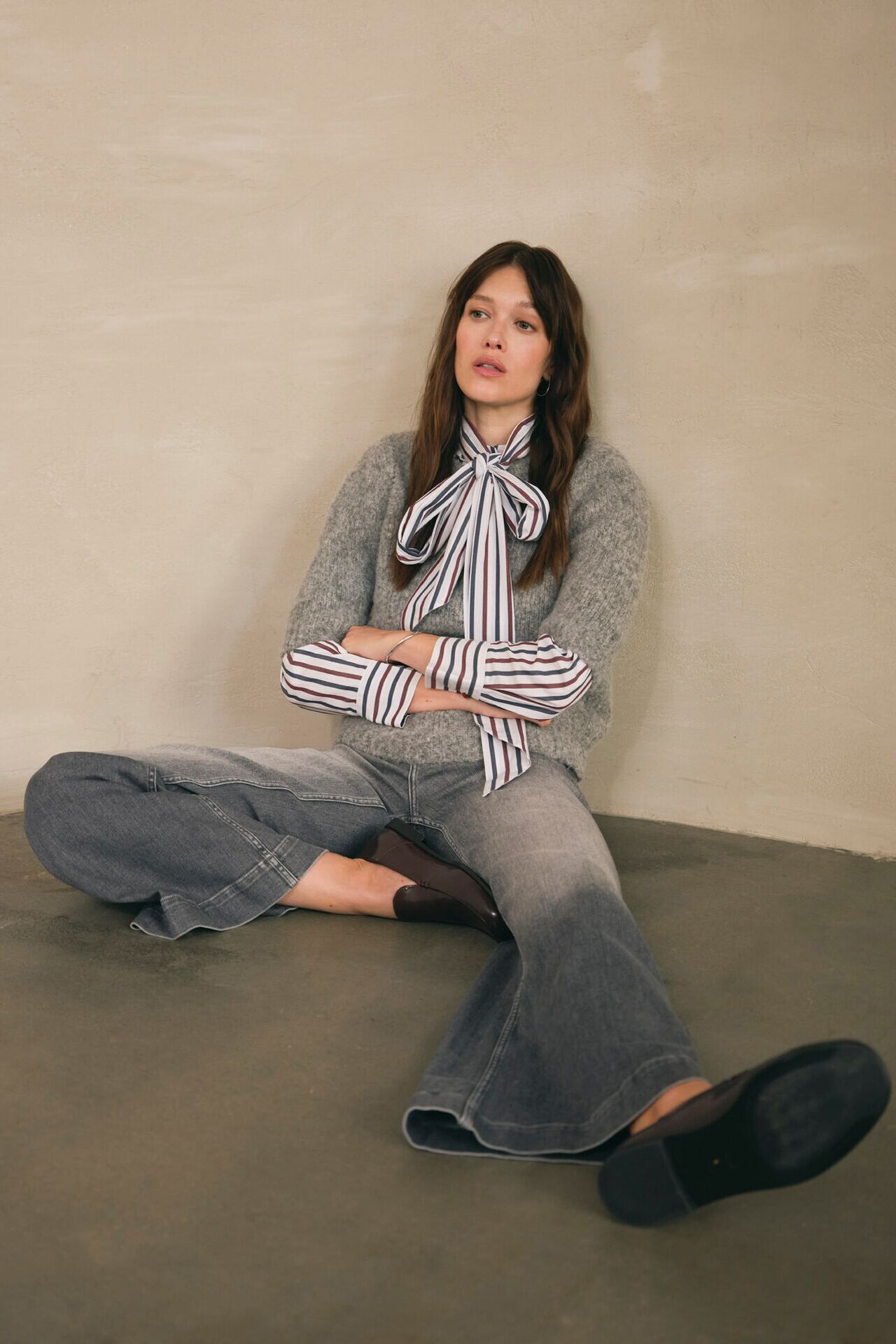 Weibliches Modell in grauer Flared-Leg Jeans, gestreifter Bluse und grauen Strickpullover sitzt auf dem Boden