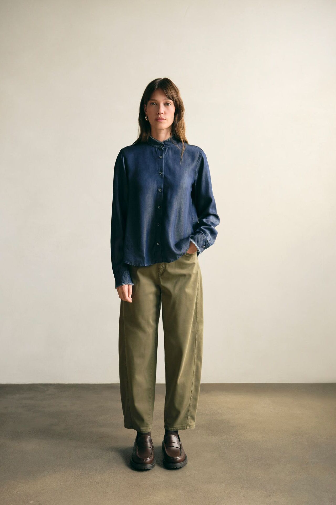 Weibliches Modell in grüner Jeans und dunkelblauer Denim-Bluse posiert mit einer Hand in der Hosentasche