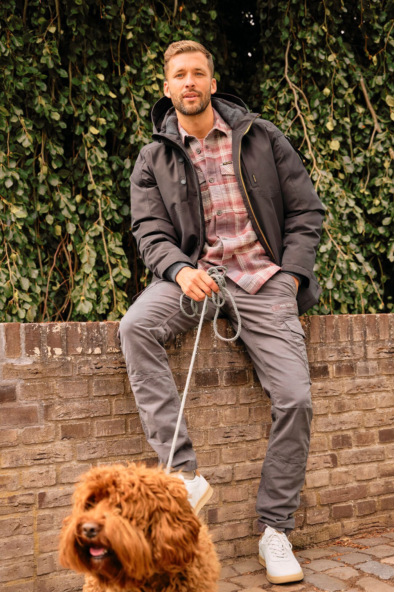 Männliches Modell in grauer Hose, karierten Hemd und dicker Jacke sitzt auf einer Mauer und hat einen Hund an der Leine