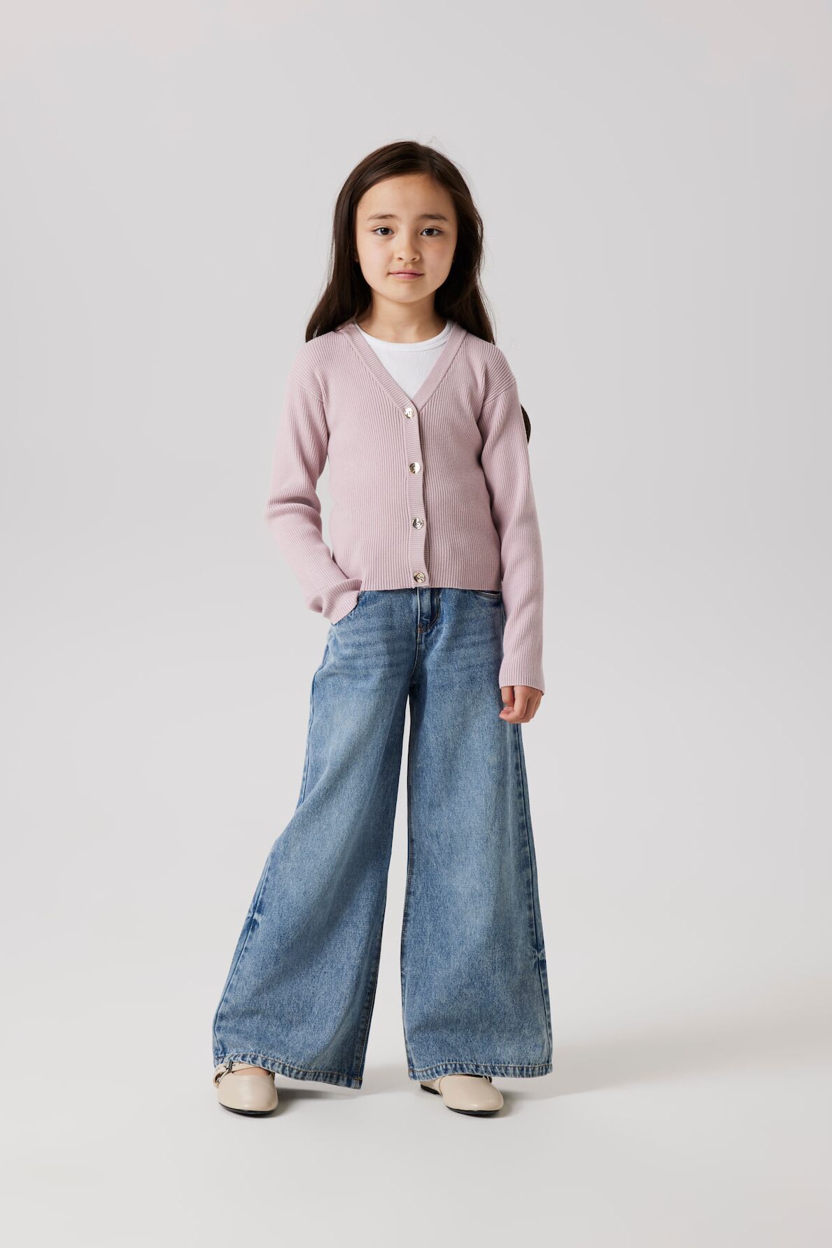 Kleines Mädchen in Wide-Leg-Jeans und rosa Cardigan von name it posiert lässig mit einer Hand in der Hosentasche
