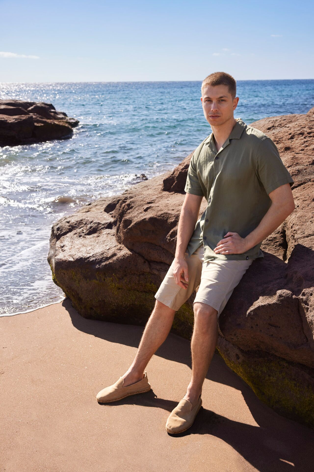 Männliches Modell in grauen Shorts und dunkelgrünen Kurzarm-Hemd lehnt sich an einen großen Felsen am Strand