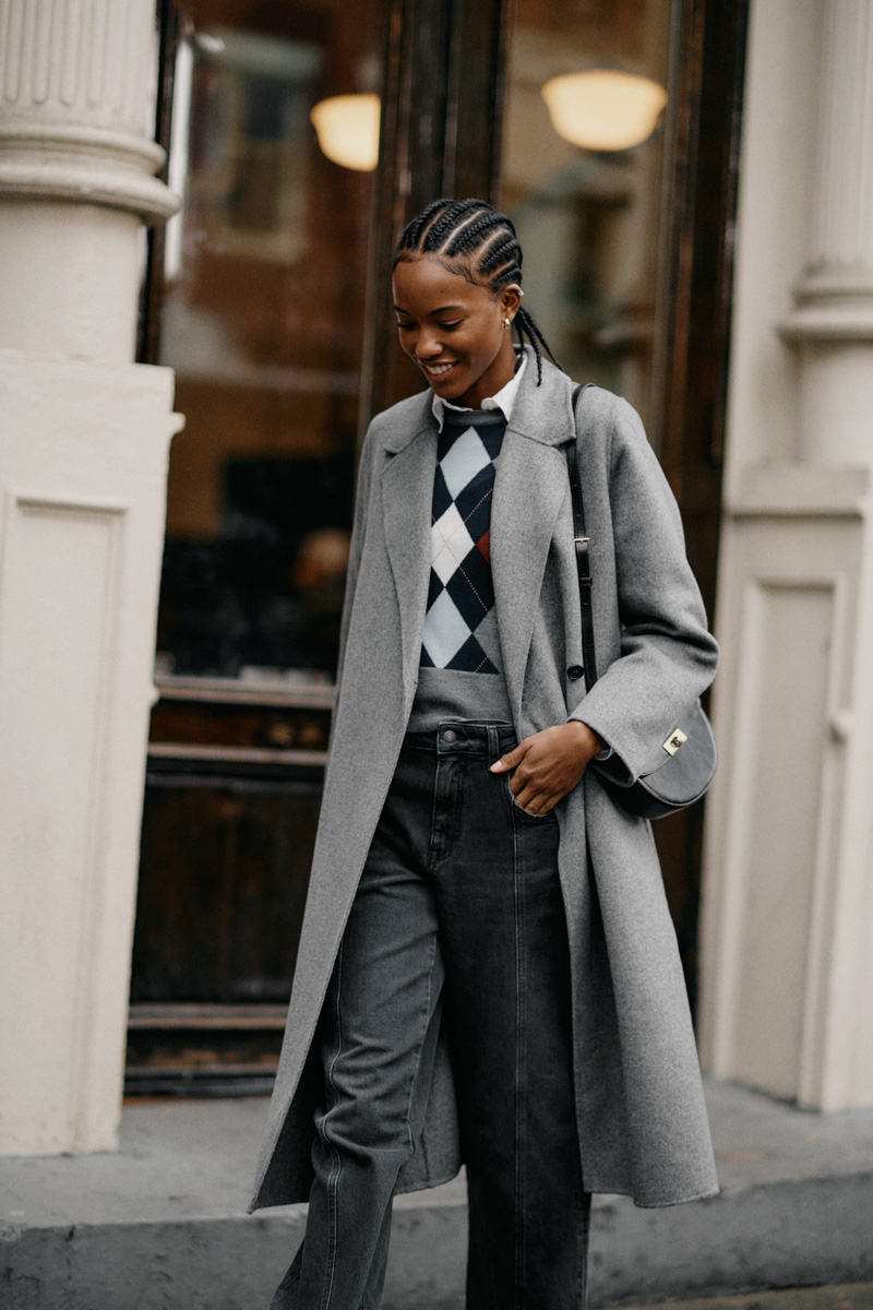 Weibliches Modell in dunkler Jeans, gemusterten Pullover und grauen Mantel von GANT läuft auf dem Gehweg