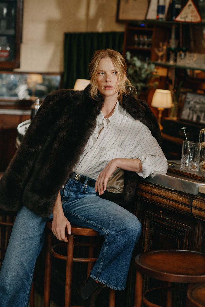 Weibliches Modell in blauer Jeans, gestreifter Bluse und übergelegter Jacke von GANT posiert an einer Bartheke