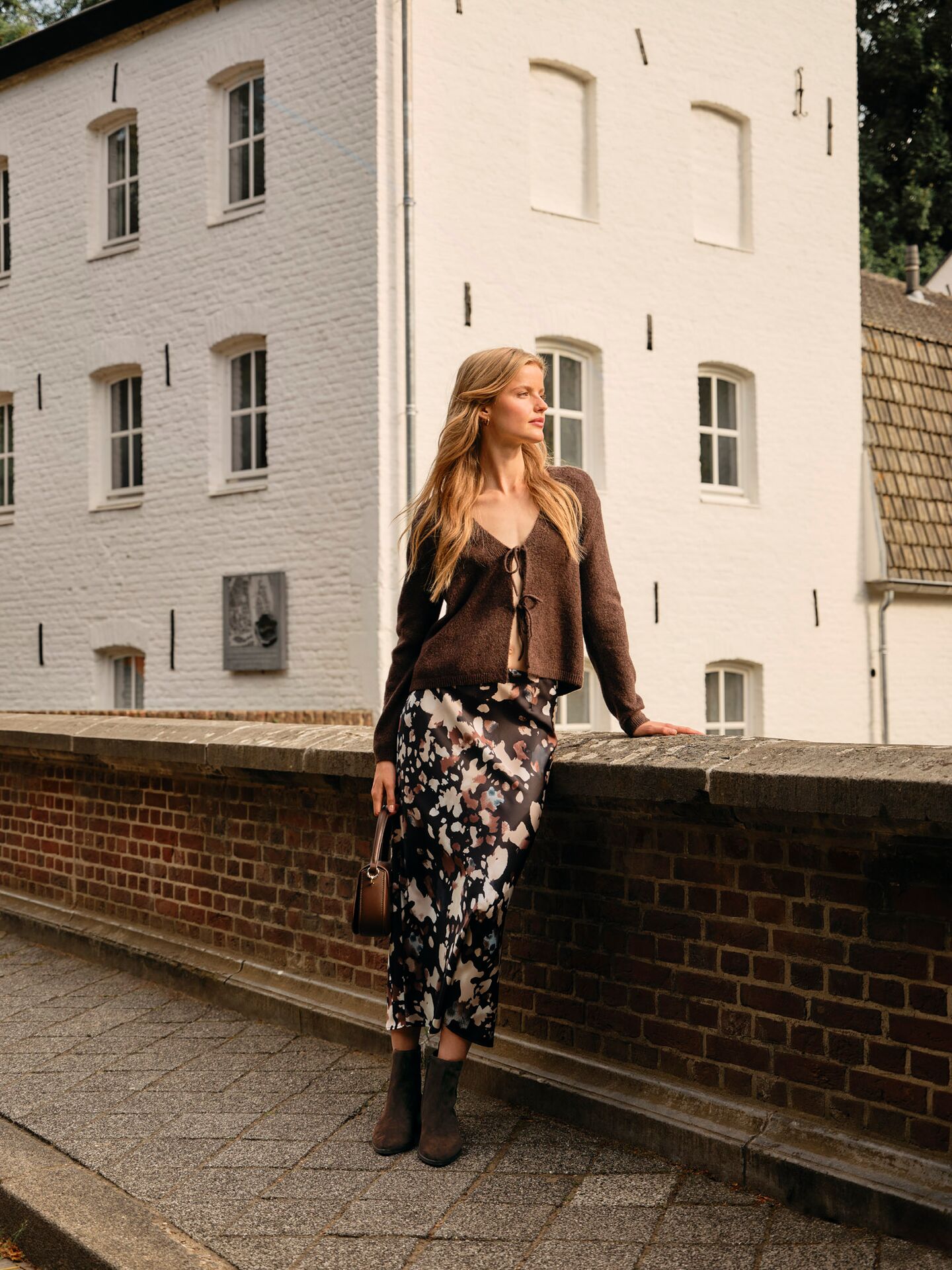 Weibliches Modell in angesagten Trend-Look aus Maxikleid und Cardigan posiert an einer Mauer