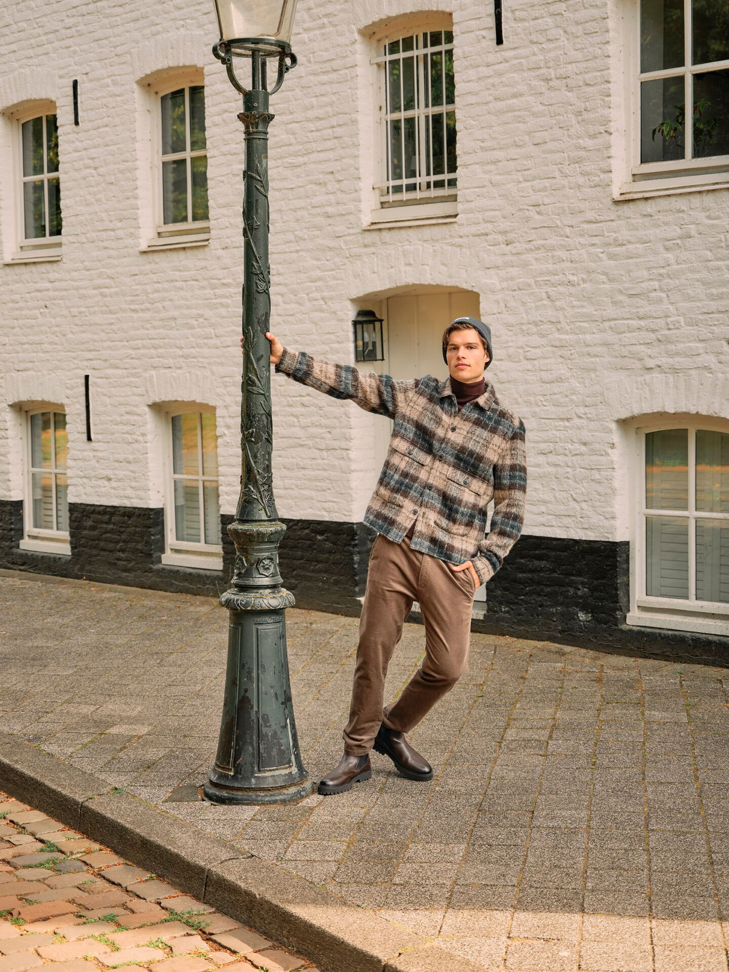 Männliches Modell in brauner Chinohose und karierten Overshirt hält sich an einer Straßenlaterne fest