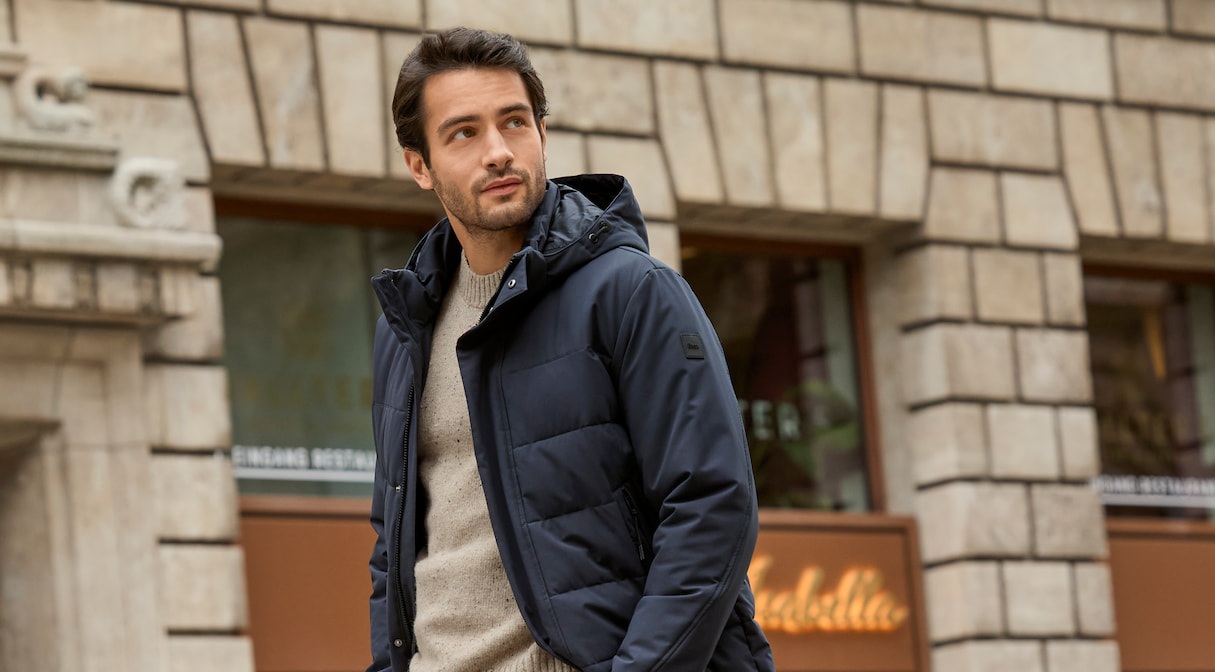 Männliches Modell in beiger Hose, beigen Pullover und dunkelblauen Steppmantel von Brax läuft entlang einer Straße